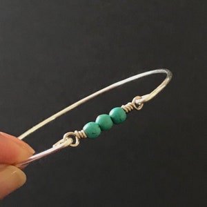 May include: A silver-toned wire bangle bracelet featuring three small, round, turquoise-coloured beads centred between two wire-wrapped loops. The metal wire is smooth and polished, creating a simple, circular band that connects to the bead section.