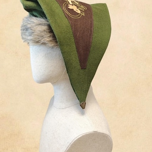 Può includere: Un cappello a punta verde con un pannello triangolare marrone caratterizzato da un emblema ricamato in oro. Il cappello presenta un bordo in pelliccia sintetica grigia sul davanti e un piccolo accento metallico triangolare sulla punta, su un manichino bianco.