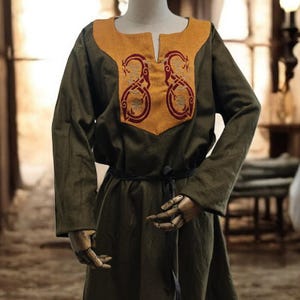 Op de afbeelding: Een olijfgroene tuniekjurk met lange mouwen en een mosterdgeel juk. Het juk is geborduurd met een rood ontwerp. Een zwarte riem sluit de taille. De jurk wordt op een mannequin getoond.