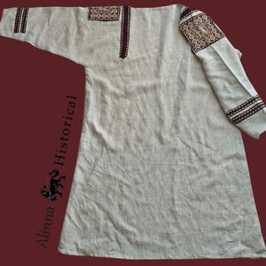Könnte beinhalten: Eine hellgraue Tunika mit langen Ärmeln. Die Tunika hat dekorative braune Stickmuster auf den Schultern und Ärmeln. Der Halsausschnitt ist V-förmig. Die Tunika ist aus einem leinenartigen Stoff gefertigt, wahrscheinlich historisch oder mittelalterlich.