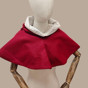 May include: A red cloak with a white-lined hood, displayed on a mannequin. The cloak has a simple design and is made of a textured fabric. The words "Alinna Historical" are printed at the top.