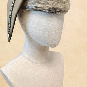 Può includere: Un cappello in feltro color marrone chiaro con una parte posteriore curva e appuntita e un'ampia fascia di pelliccia sintetica marrone e crema sulla fronte. Il bordo del cappello presenta una decorazione nera con un motivo a catena dorata.
