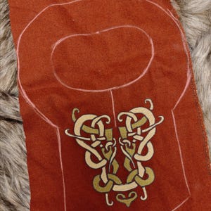 Könnte beinhalten: Rostfarbener Stoff mit einem keltischen Knotenmuster, gold und grün bestickt. Der Stoff hat eine gezeichnete Umrisslinie, die auf ein Kleidungsstückmuster hindeutet. Das Alinna Historical Logo ist unten rechts zu sehen.