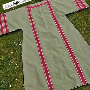 Puede incluir: Una túnica verde oliva con ribetes burdeos y costuras decorativas blancas. La túnica tiene mangas cortas y está sobre una superficie cubierta de hierba. El texto "Alinna Historical" está en la esquina superior izquierda.