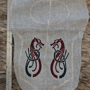 Peut inclure: Une pièce de tissu en lin gris clair ornée de deux motifs de dragons symétriques rouge foncé et vert avec des entrelacs complexes. Une règle métallique indique une longueur de 50 cm et une largeur de 30 cm. Le texte Alinna Historical est visible.