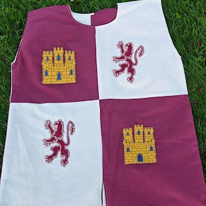 May include: A sleeveless, medieval-style tunic in a four-square design. The tunic features alternating white and burgundy panels. Each panel has an embroidered emblem: a castle and a lion. The text "Alinna Historical" is printed at the bottom.