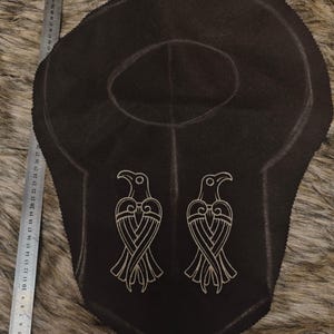 May include: A dark brown fabric piece with a rounded neck opening and two embroidered bird figures in a light tan thread. A metal ruler is placed along the left side and bottom edge, showing the item measures approximately 30 cm in width.