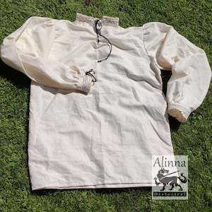 Può includere: Una camicia di lino bianca con colletto alto e maniche lunghe. La camicia ha un laccio in corda nero al collo e un bordo nero attorno al colletto e ai polsini.