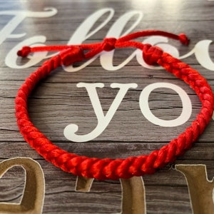 May include: A bright red braided bracelet with an adjustable knot fastening. The bracelet is made of a thick, woven material and is displayed on a wooden surface with the words "Follow Your" in the background.