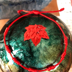 May include: Red string bracelet with a red maple leaf charm. The bracelet is tied with knots.