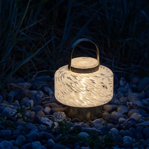 May include: A white glass lantern with a metal handle and a swirling pattern. The lantern is lit and casting a warm glow. It is sitting on a bed of small, gray stones.