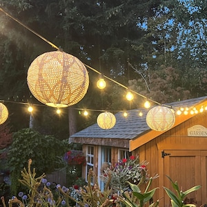 May include: Outdoor scene with string lights and decorative globe lanterns. A wooden shed with a "Garden" sign is in the background. The lanterns are lit, creating a warm, inviting ambiance for a backyard or garden setting.