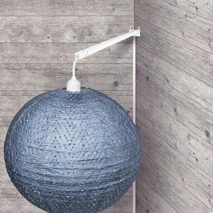 May include: A blue paper lantern with a textured surface and small perforations hangs from a white, adjustable arm. The spherical lamp is suspended against a wooden wall, creating a modern lighting fixture.