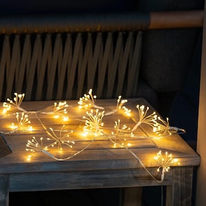 May include: A string of LED fairy lights featuring multiple starburst-shaped clusters of warm white bulbs connected by thin, flexible silver-toned wire. Each cluster radiates outward with small, glowing points of light.