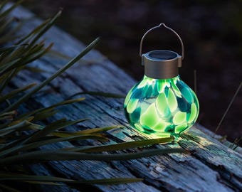 May include: A solar-powered, green glass lantern with a silver handle and rim. The lantern has a rounded shape and is filled with glowing green accents. It sits on a weathered wooden surface, with green foliage in the foreground.