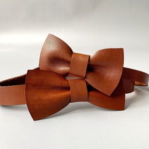 Könnte beinhalten: Zwei braune Leder-Fliegen, eine größere und eine kleinere, sind durch ein braunes Lederband verbunden.