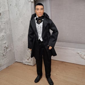 May include: A doll in a black formal suit, featuring a long coat, trousers, and shoes. The outfit includes a white patterned waistcoat, a black bow tie, and a decorative brooch. The doll has dark hair. The doll is standing in a room.