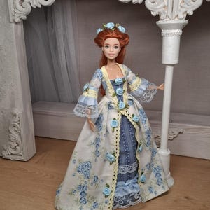 May include: A doll wearing a historical-style gown. The dress is white with a blue floral pattern and blue rose accents. The sleeves are trimmed with lace and yellow ribbon. The doll has red hair and a matching floral headpiece.