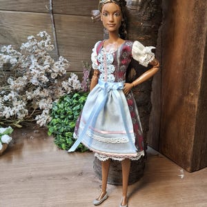 Könnte beinhalten: Puppe in einem traditionellen deutschen Dirndl-Kleid. Das Kleid hat ein Blumenmuster mit roten und weißen Akzenten, eine hellblaue Schürze und weiße Spitzenbesätze. Die Puppe hat braune Haut, braune Haare und silberne Schuhe.