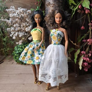 May include: Two fashion dolls are displayed. One doll wears a yellow top with a green trim and a floral skirt. The other doll is in a white lace dress with a floral bodice. Both dolls have brown skin and are wearing gold heels.