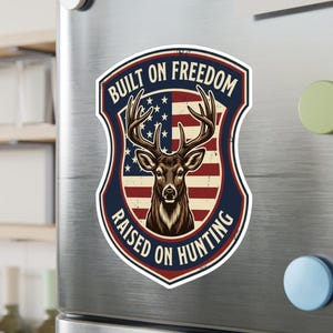 Puede incluir: Un imán en forma de escudo con una ilustración de ciervo y el texto "BUILT ON FREEDOM RAISED ON HUNTING". El diseño presenta un ciervo con cuernos sobre un fondo de la bandera estadounidense, enmarcado por un borde azul marino.