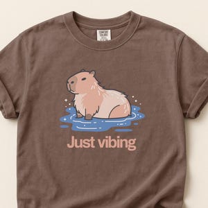 Puede incluir: Camiseta marrón con un gráfico de un carpincho en el agua, con el texto rosa "Just vibing". El carpincho es marrón claro y rosa, y el agua es azul. La camiseta tiene mangas enrolladas.