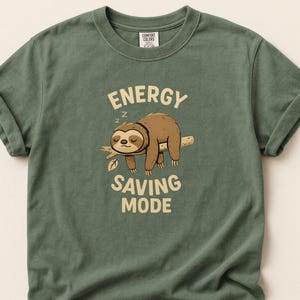 Puede incluir: Camiseta verde con mangas enrolladas. Presenta un perezoso de dibujos animados durmiendo en una rama, con el texto "ENERGY SAVING MODE" arriba y abajo. La camiseta tiene cuello redondo y una etiqueta Comfort Colors.