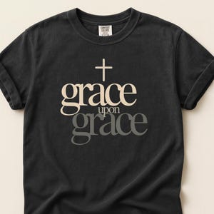 Puede incluir: Camiseta negra con mangas enrolladas. La camiseta presenta las palabras "grace upon grace" en un diseño en capas, con una cruz encima de la palabra "grace". La camiseta está hecha de un material suave.