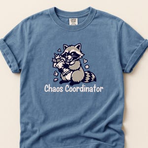 Puede incluir: Camiseta azul con un dibujo de un mapache de dibujos animados sosteniendo papeles y rodeado de escombros. El texto "Chaos Coordinator" está impreso debajo. La camiseta tiene mangas enrolladas y es de material suave.