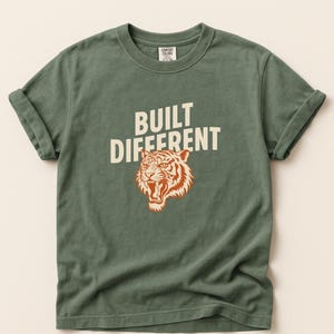 Puede incluir: Camiseta verde con mangas enrolladas. La camiseta presenta el texto "BUILT DIFFERENT" sobre un gráfico de cabeza de tigre en colores naranja y crema. La camiseta está hecha de un material suave y cómodo.