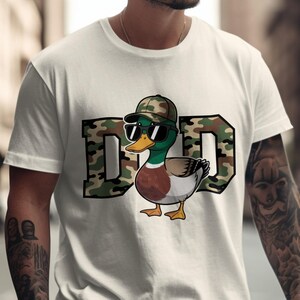 May include: White t-shirt with a graphic of a duck wearing sunglasses and a camouflage hat. The word "DAD" is printed in camouflage letters. The duck has green, brown, and white feathers.