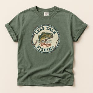 Puede incluir: Camiseta verde oliva con mangas enrolladas. En la parte delantera, un gráfico circular con un pez saltando del agua y el texto "LESS TALK MORE FISHING". El gráfico utiliza tonos marrones, verdes y azules.