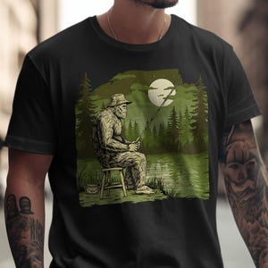 Puede incluir: Camiseta negra con un gráfico de Bigfoot pescando junto a un lago bajo la luna llena. La ilustración es en tonos verdes, beige y negro. El Bigfoot está sentado en un taburete, sosteniendo una caña de pescar. El diseño incluye árboles y un cubo.