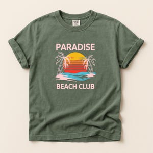 Puede incluir: Camiseta verde oliva con mangas enrolladas. La parte delantera presenta un gráfico de una puesta de sol sobre el océano con palmeras y las palabras "PARADISE BEACH CLUB" en rosa. La camiseta está hecha de un material suave y cómodo.