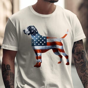 May include: Cream-colored t-shirt featuring a graphic of a dog with the American flag design. The dog's body is red and white striped, with a blue head and stars. The shirt is a casual style.