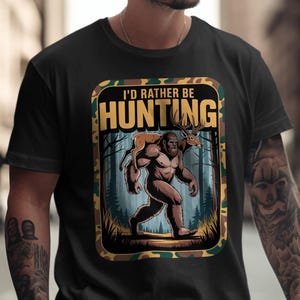 Puede incluir: Camiseta negra con un gráfico que presenta a Bigfoot llevando un ciervo, con el texto "I'd Rather Be Hunting". El diseño está enmarcado con un borde de camuflaje, y el fondo muestra una escena de bosque.