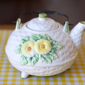 May include: White ceramic teapot with a woven design and floral accents. The teapot has a lid and a handle with a metal wire.