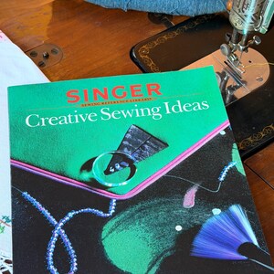 May include: A Singer Sewing Reference Library book titled Creative Sewing Ideas with a green and black cover featuring a graphic of a blue beaded necklace, a triangular fabric piece, and a blue fan-shaped brush, resting near a sewing machine.