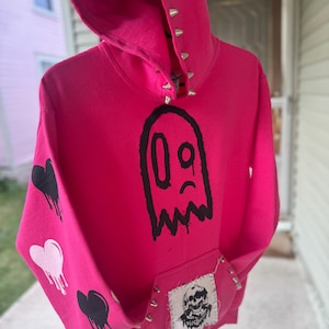 Puede incluir: Una sudadera con capucha de color rosa brillante que presenta un gran gráfico de fantasma negro en el pecho, tachuelas metálicas plateadas en el borde de la capucha y corazones negros y rosa claro goteando en la manga. Un parche con calavera está cosido.