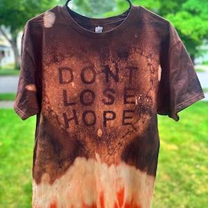 May include: A short-sleeve crew neck t-shirt with a dark brown and orange bleach-dyed pattern. The center of the shirt features the text "DONT LOSE HOPE" in a dark brown font, set against a lighter, heart-shaped bleached area on the fabric.