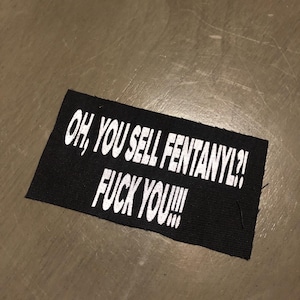 Könnte beinhalten: Ein schwarzer rechteckiger Aufnäher mit weißem Text, der lautet: "OH, YOU SELL FENTANYL?! FUCK YOU!!!". Der Aufnäher ist wahrscheinlich aus Stoff und liegt auf einer neutralfarbenen Oberfläche.