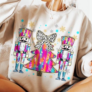 May include: A white sweatshirt with a Christmas tree design featuring two nutcracker figures on either side of the tree. The tree is decorated with a large black and white polka dot bow and colorful abstract paint.