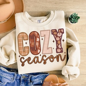 May include: A cream-colored sweatshirt with the text "Cozy Season" in brown, pink, and orange letters with a plaid and floral pattern. The letters are outlined in black and have a glittery effect.