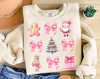 May include: A cream-colored sweatshirt with pink Christmas-themed iron-on patches. The patches include a gingerbread man, Santa Claus, a Christmas tree, a bow, a nutcracker, and a gift box.