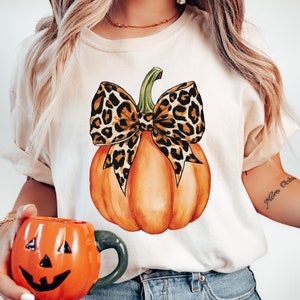 May include: A white t-shirt with a graphic of an orange pumpkin with a leopard print bow. The pumpkin is centered on the shirt.