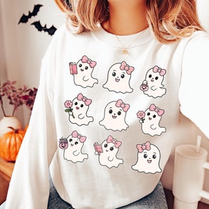May include: White sweatshirt with a graphic print of nine cute cartoon ghosts wearing pink bows. Each ghost has a different activity, such as reading a book, holding a flower, or carrying a pumpkin.
