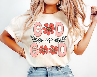May include: Cream-colored t-shirt with the text "GOD is GOOD" in a pink, retro font. The letters "O" are replaced with pink flowers. Green leaves and stems are between the words. The model is wearing light blue jeans.