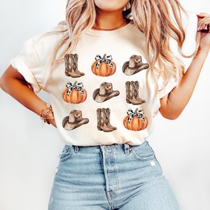 May include: A white t-shirt with a pattern of brown cowboy hats, boots, and orange pumpkins with black and white polka dot bows.
