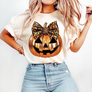 May include: A white t-shirt with a gold glitter pumpkin with a leopard print bow. The pumpkin has a jack-o'-lantern face.