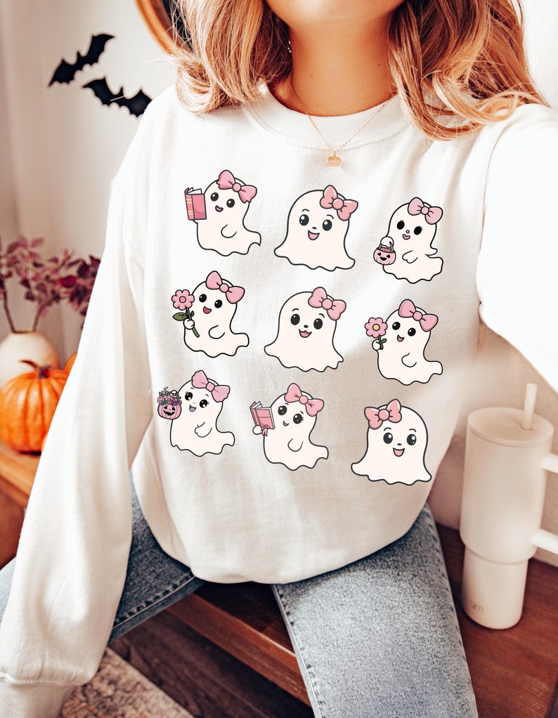 May include: White sweatshirt with a graphic print of nine cute cartoon ghosts wearing pink bows. Each ghost has a different activity, such as reading a book, holding a flower, or carrying a pumpkin.